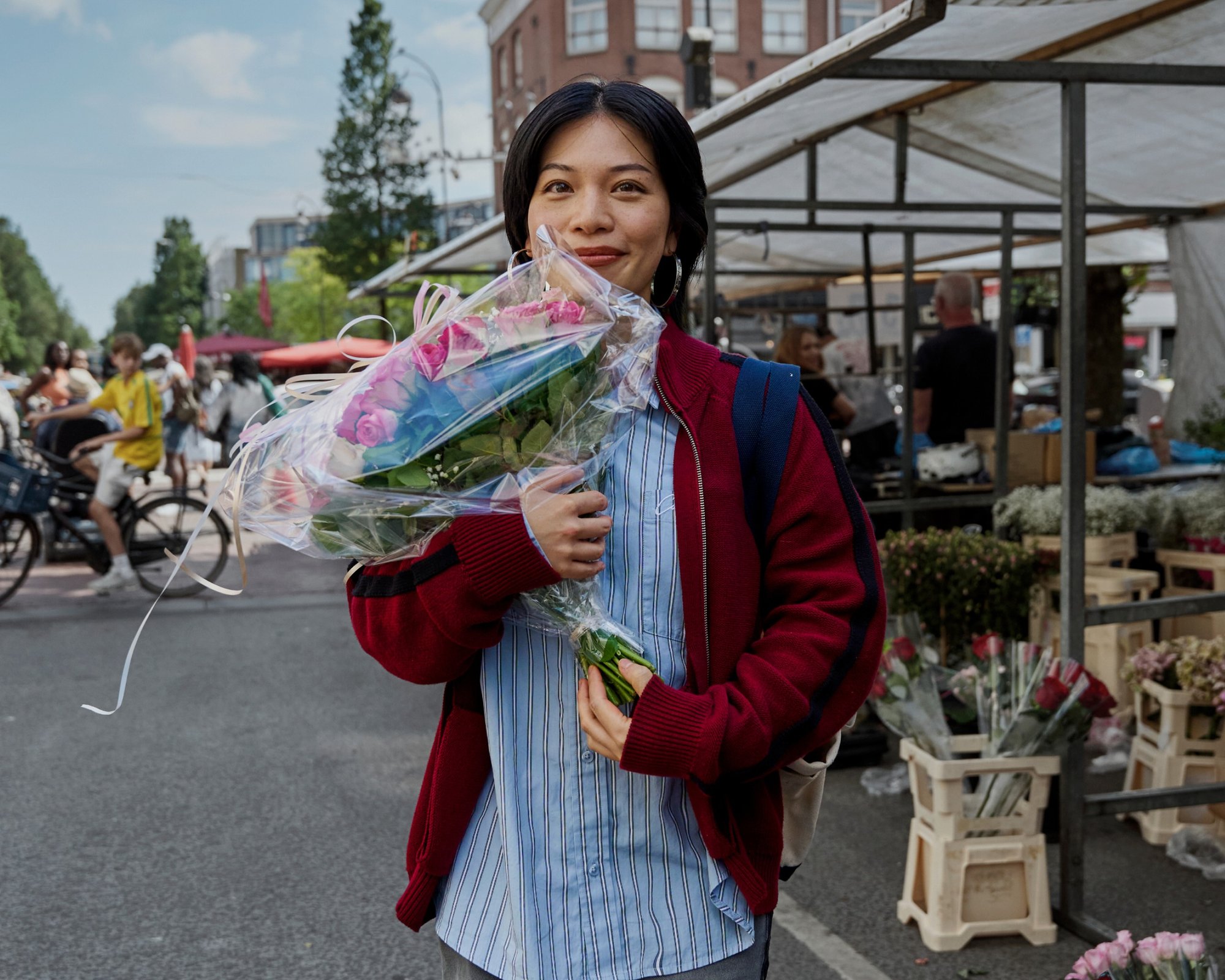 Un bouquet de roses roses, emballé dans du plastique transparent avec un ruban blanc. Une personne porte un cardigan rouge par-dessus une chemise à rayures bleues, tenant uneplus petite fleur.