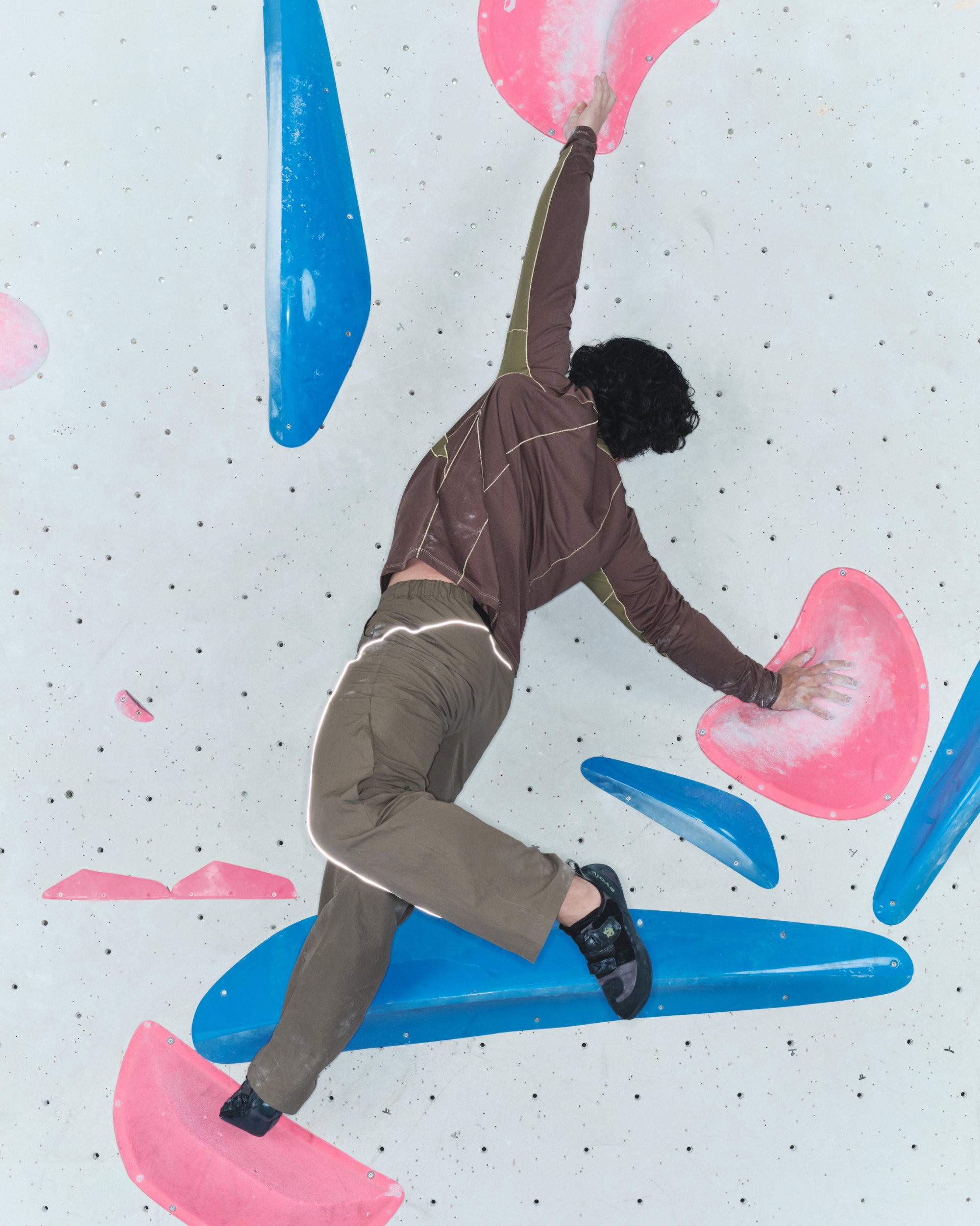 Persona vestida con ropa marrón escalando una pared interior, agarrándose de grandes presas de escalada rosas y azules.