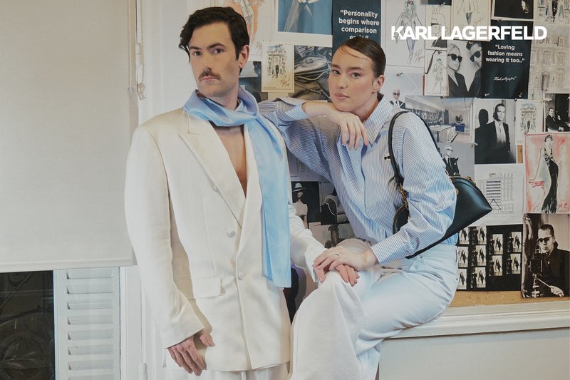 Man in white suit and blue scarf stands next to woman in blue striped shirt and white pants, posing in front of a Karl Lagerfeld photo collage.