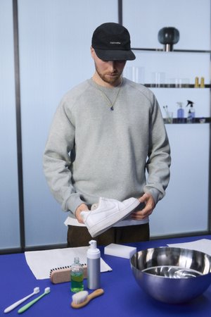 A person in a gray sweatshirt holds a white sneaker, surrounded by cleaning supplies on a blue table and a modern, minimalist background.