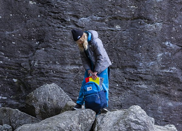 Blå ryggsekk med flere rom, plassert foran en steinete bakgrunn. Brukeren har på seg en grå jakke og blå bukser, og sitter på huk.