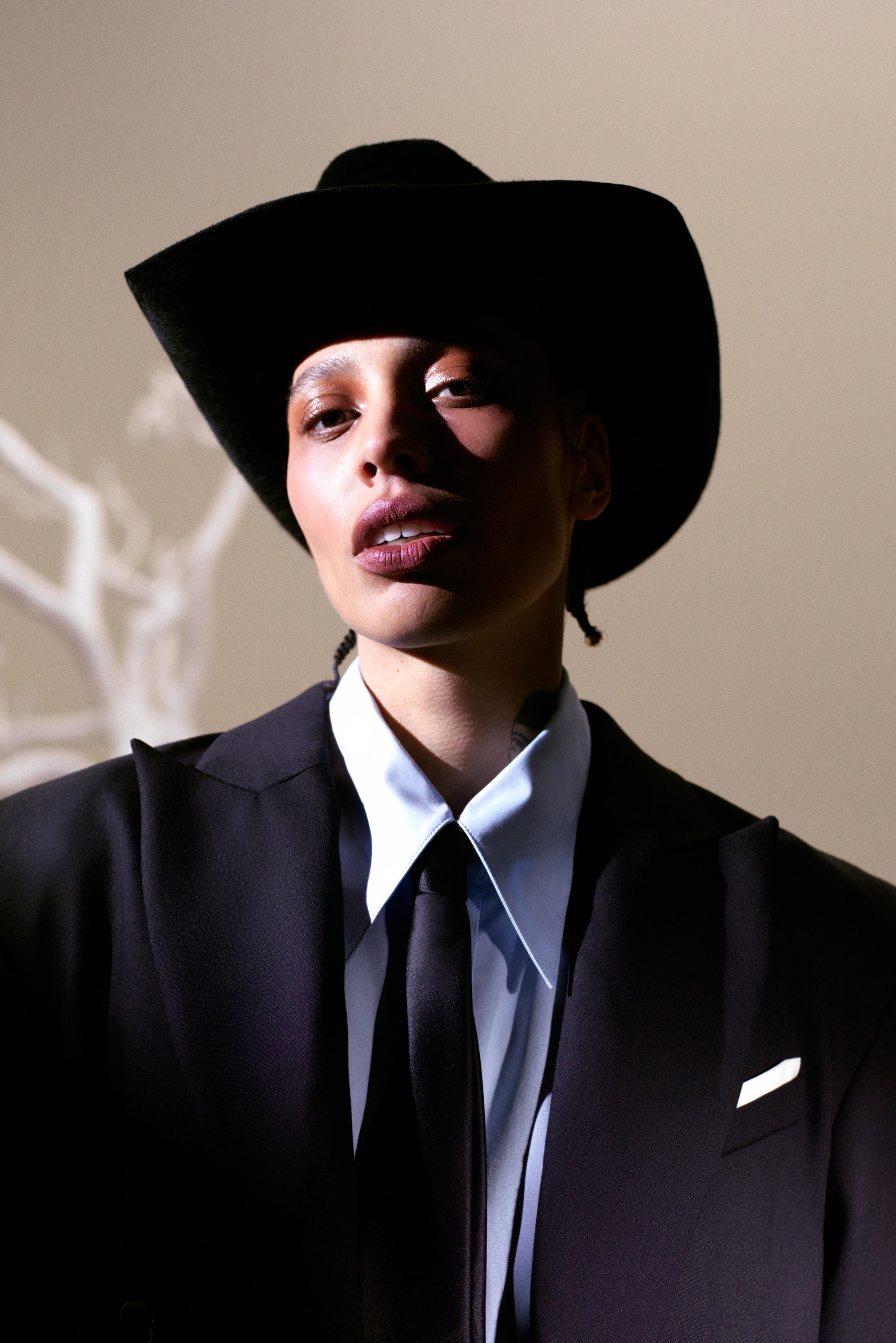A person with braided hair wears a black cowboy hat and a sharp suit, confidently facing the camera against a softly lit background.