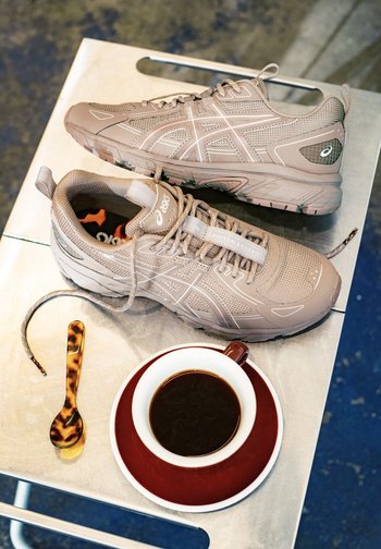 Beige athletic shoes rest on a table beside a cup of black coffee and a small spoon. The setting features a textured surface.