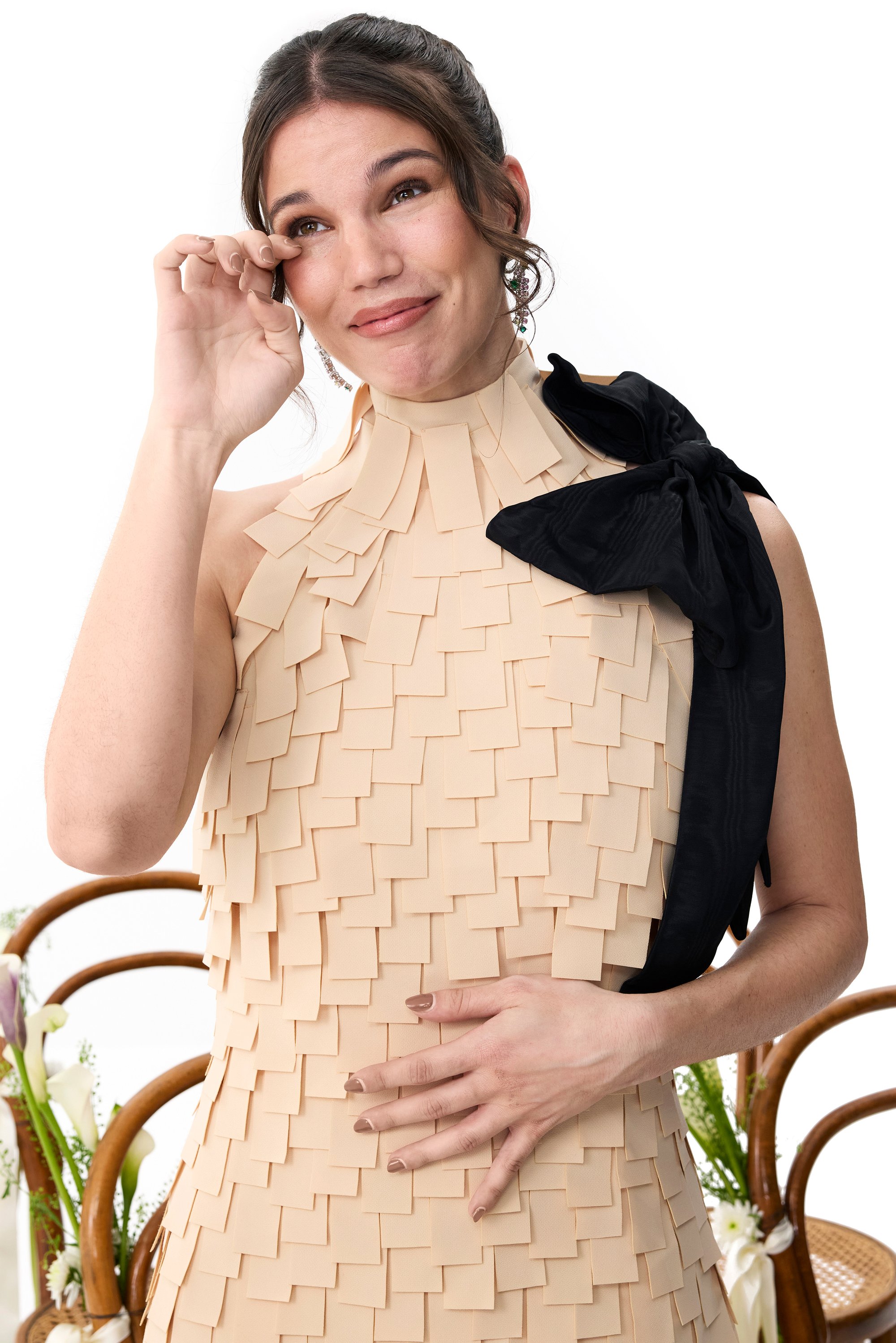 Mujer sonriente con un vestido beige texturizado se seca una lágrima, mostrando emoción. Reposa una mano sobre su estómago, enmarcada por flores y sillas.