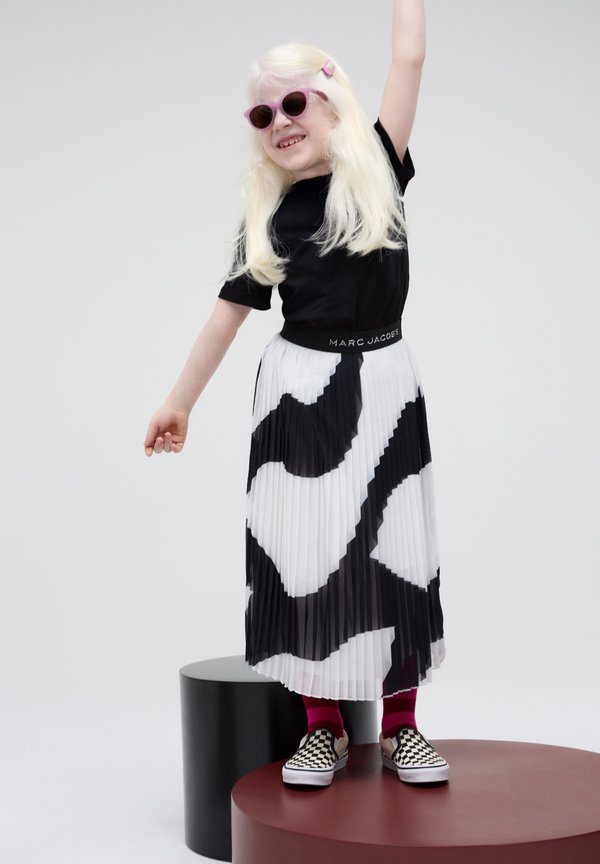 T-shirt nera, gonna plissettata bianca con motivo astratto nero, occhiali da sole rosa e scarpe slip-on a scacchi.