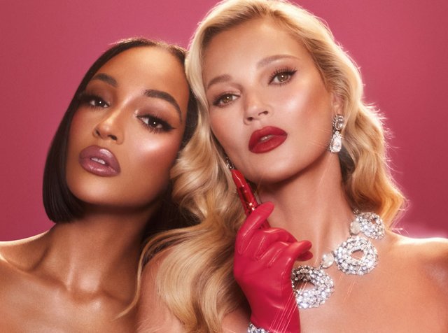 Two women pose close together, one with a bob haircut and the other with long, wavy hair, both featuring glamorous makeup and jewelry.