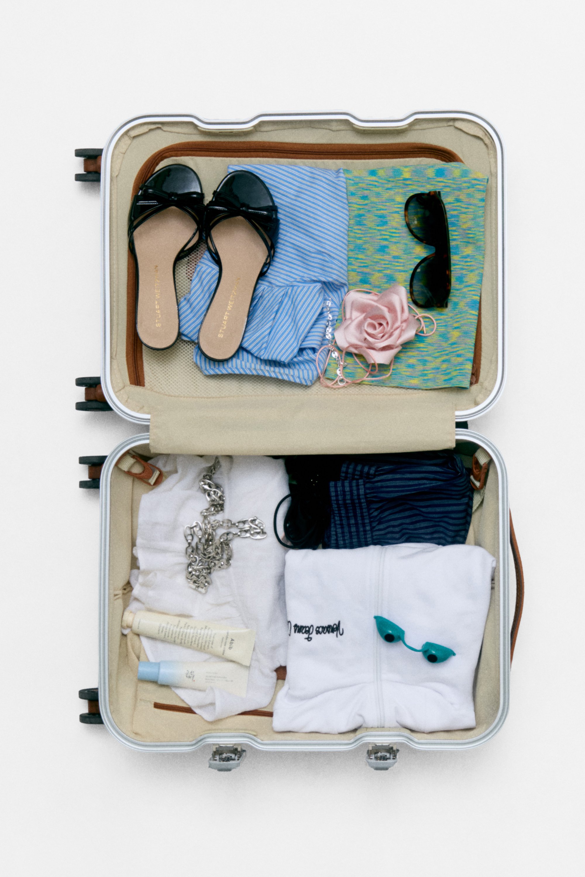 Una maleta llena de artículos esenciales para el verano: sandalias negras, ropa, gafas de sol, una rosa rosa, artículos de tocador y una sudadera blanca.