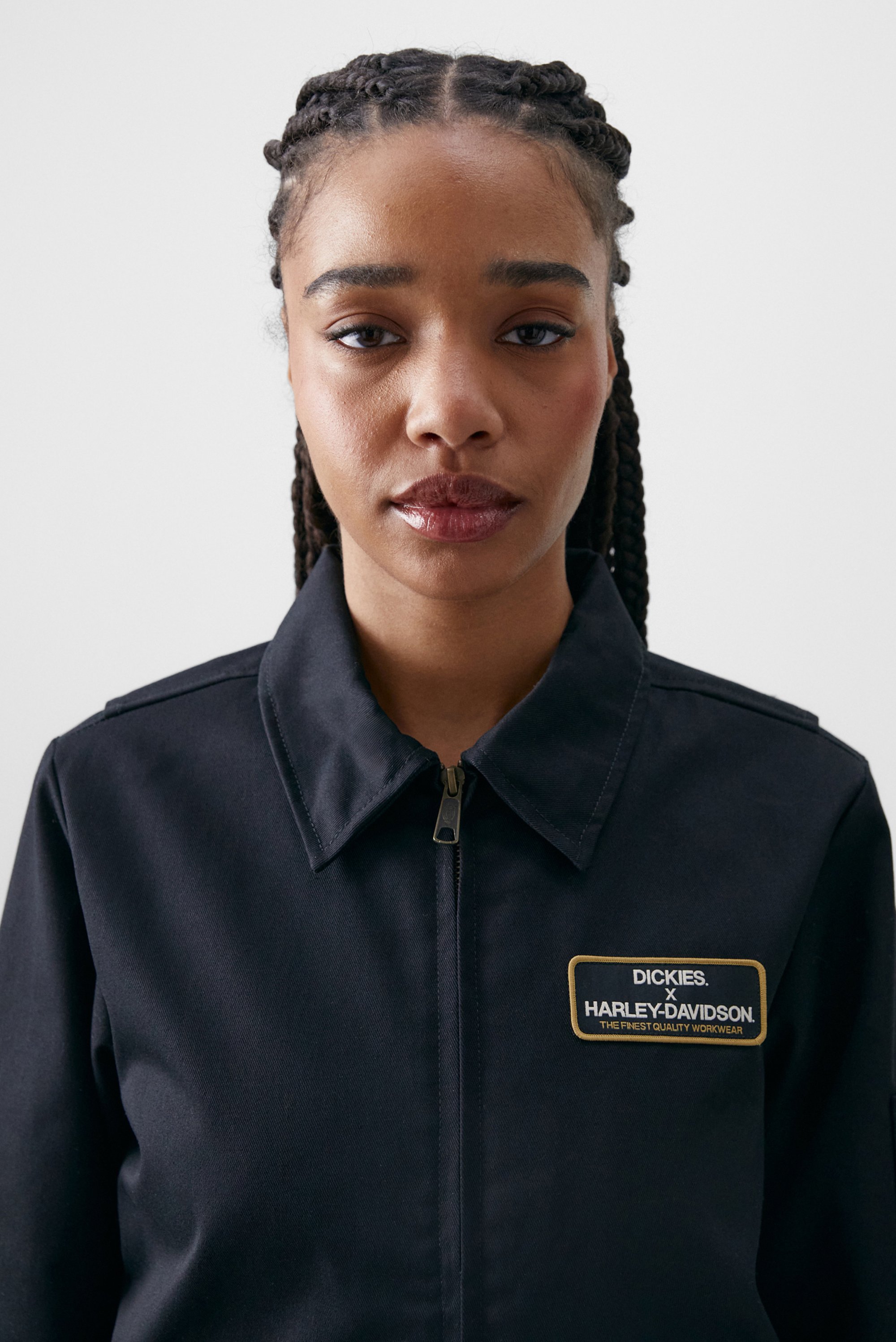 Een jonge vrouw met vlecht haar draagt een marineblauwe werkjack met een Dickies x Harley-Davidson logo. Ze kijkt recht in de camera.
