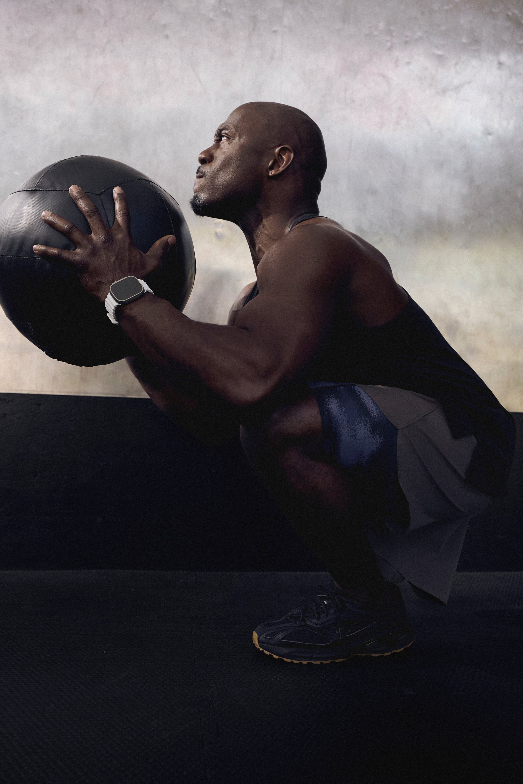 A man crouches while holding a large black medicine ball, focused and intense, in a dimly lit gym setting.