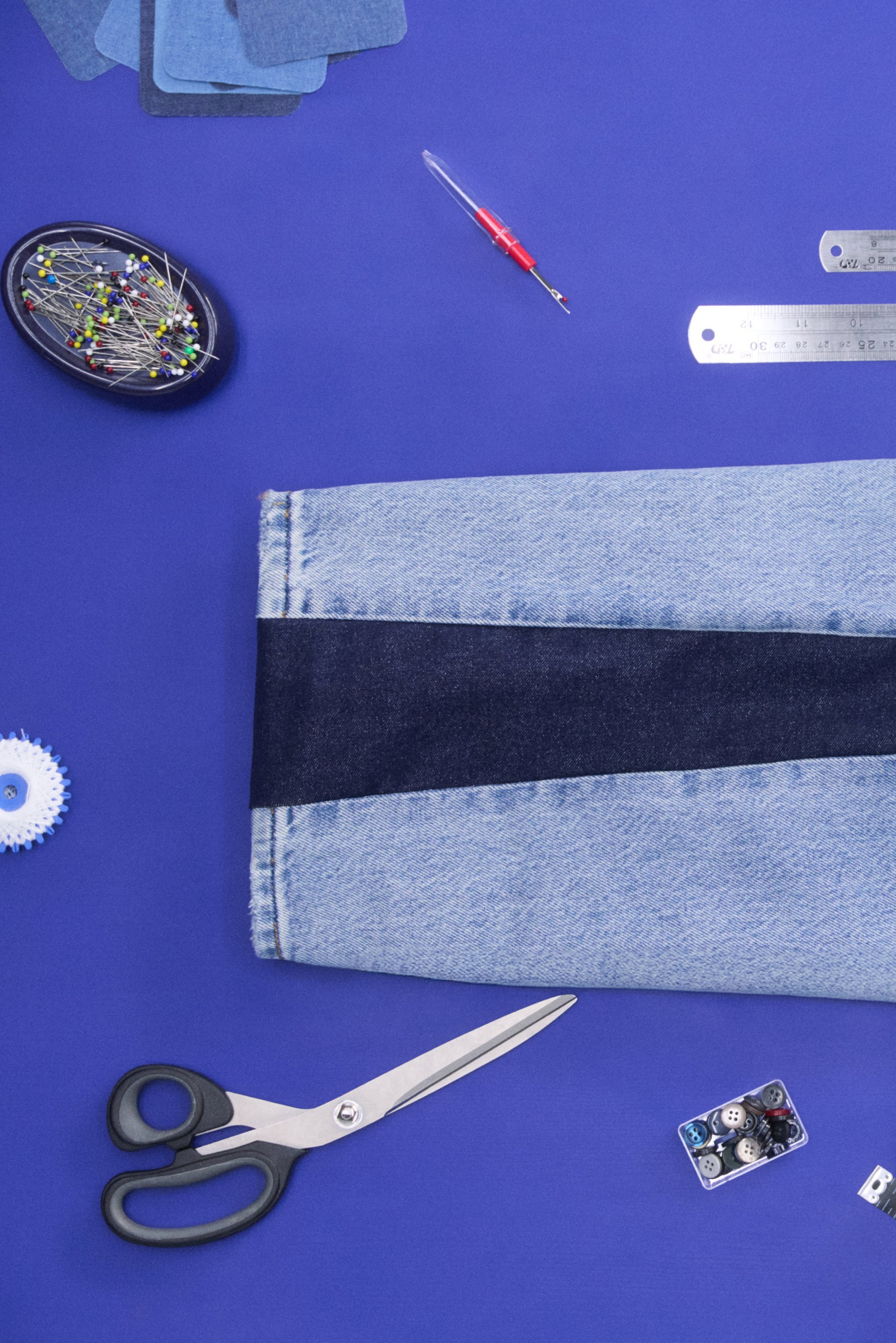 Schaar, stukjes stof, spelden en knopen zijn gerangschikt op een paarse ondergrond, wat naaitools en materialen toont voor een denimproject.