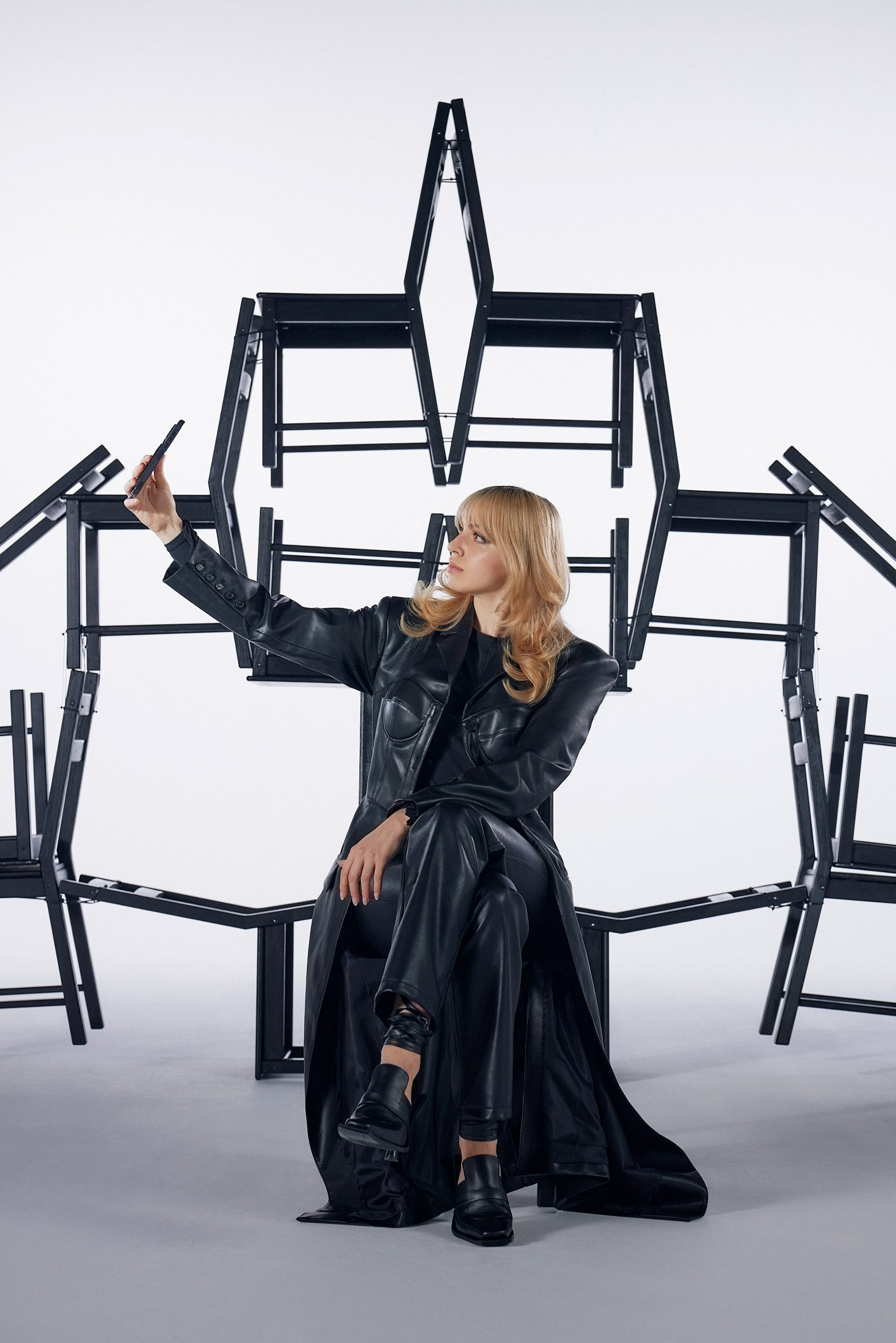 Una mujer con un elegante atuendo negro posa para un selfie, sentada entre modernas sillas negras dispuestas en una formación abstracta.