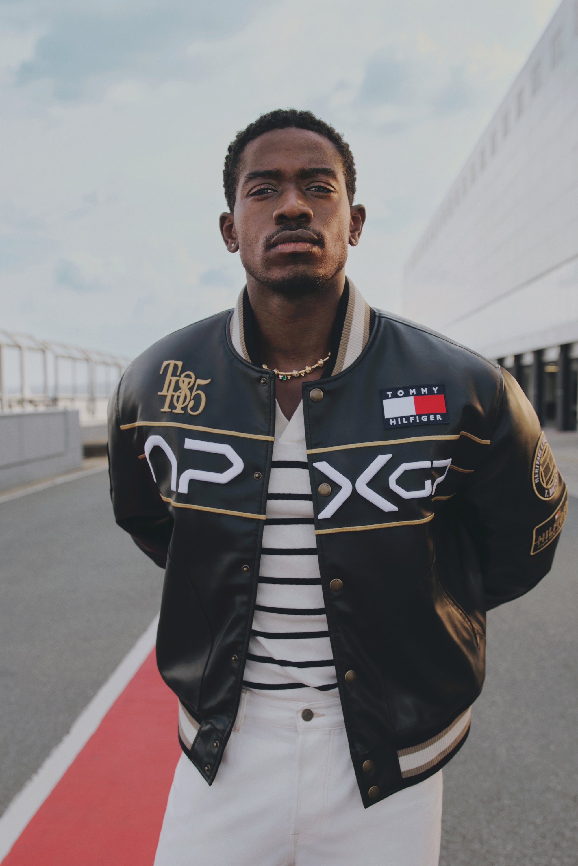 A man stands confidently, wearing a black varsity jacket with logos, a striped shirt, and white pants, against an urban backdrop.