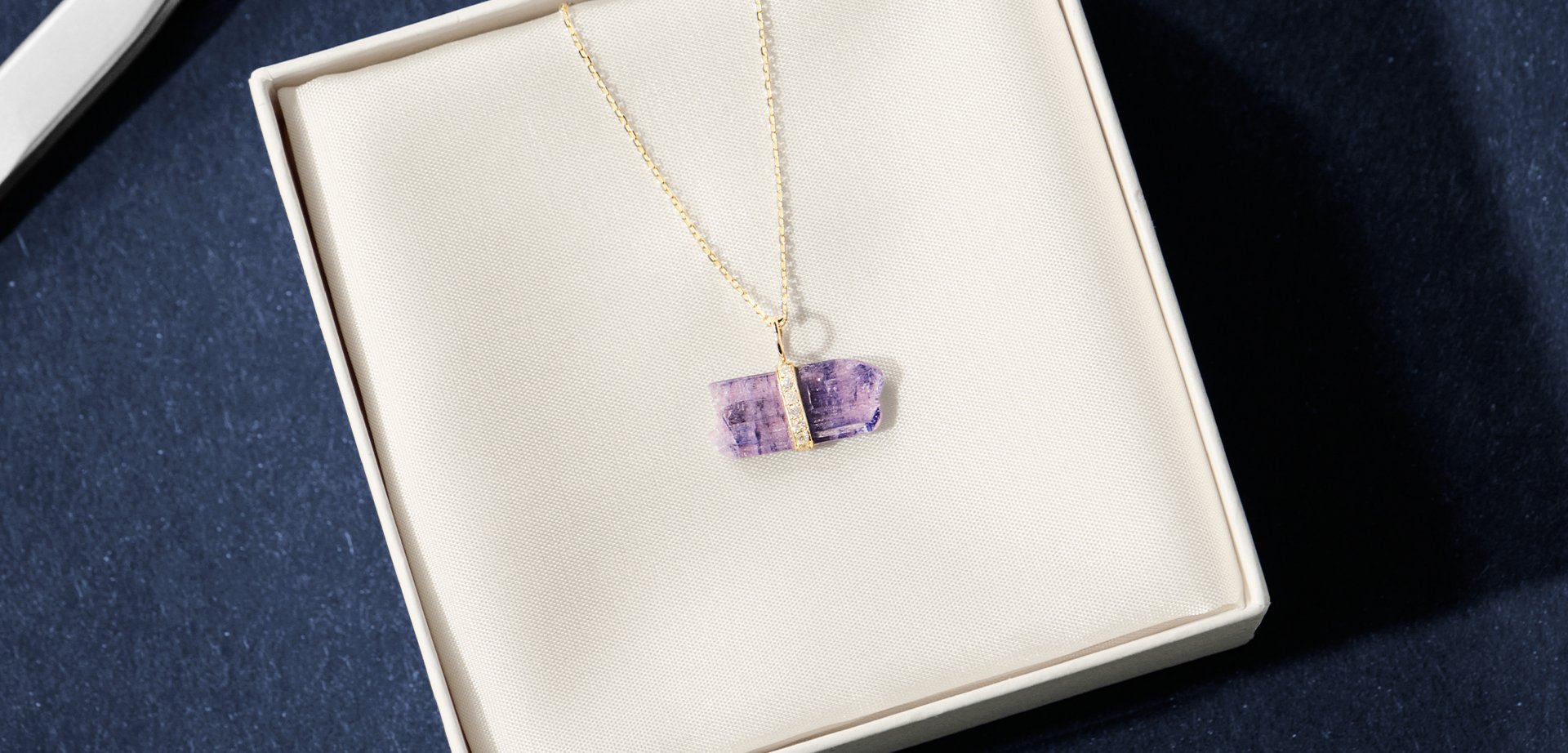The gold necklace showcases a horizontally set rectangular purple gemstone pendant adorned with a textured gold accent, presented in a cream-coloured box.