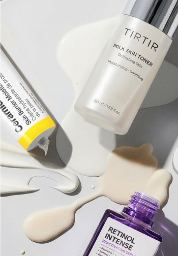 TIRTIR Tonique au lait dans un flacon givré, Crème hydratante pour la barrière cutanée CeraVe dans un tube à embout jaune, et sérum rétinol intense dans un flacon violet, le tout sur une surface blanche avec des produits renversés.