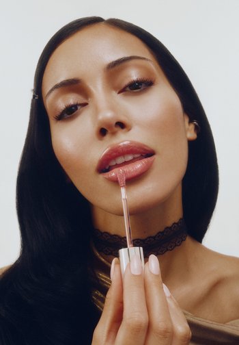 Läppglans i ett metalliskt silverrör med en svampapplikator, applicerat på fylliga läppar. Modellen har långt mörkt hår och bär en spetsig choker.