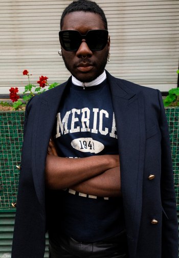 Person in Sonnenbrille und einer marineblauen Blazer steht mit verschränkten Armen, trägt einen grafischen Sweatshirt, mit roten Blumen und Grün im Hintergrund.
