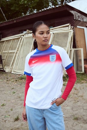 Joven mujer que lleva una camiseta de fútbol de Inglaterra con mangas rojas, de pie al aire libre cerca de estructuras de madera, mirando con confianza hacia un lado.