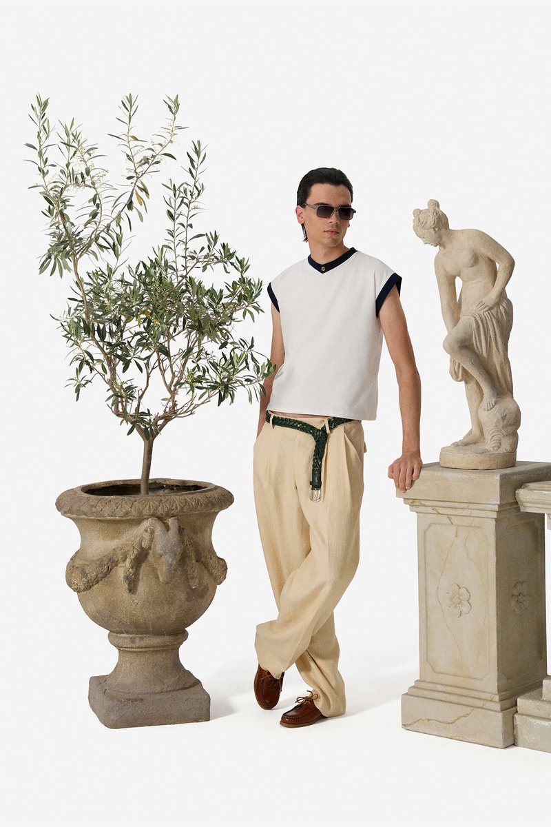 Man wearing sunglasses, sleeveless white top, beige pants, and brown shoes standing between a potted plant and a stone statue on pedestal.