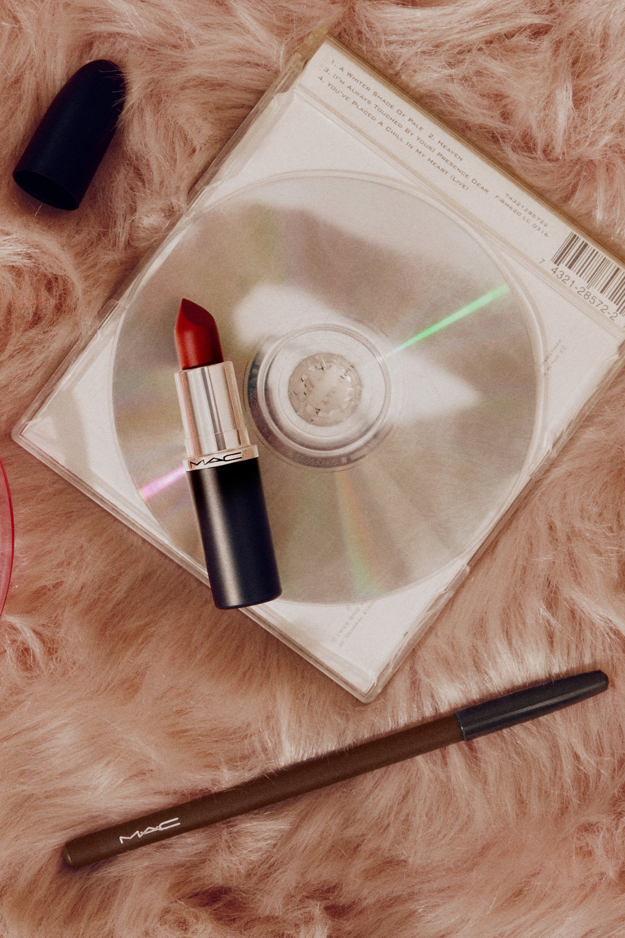 Lápiz labial rojo MAC con estuche plateado y negro, funda de CD transparente y lápiz MAC marrón sobre fondo esponjoso.