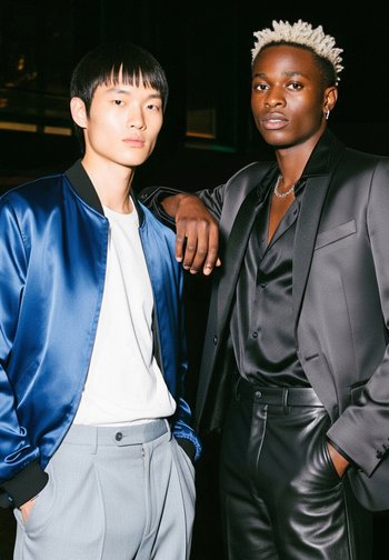 Due modelli posano insieme. Uno indossa una giacca bomber blu lucido sopra una camicia bianca, mentre l'altro sfoggia una camicia di raso nera e pantaloni di pelle.