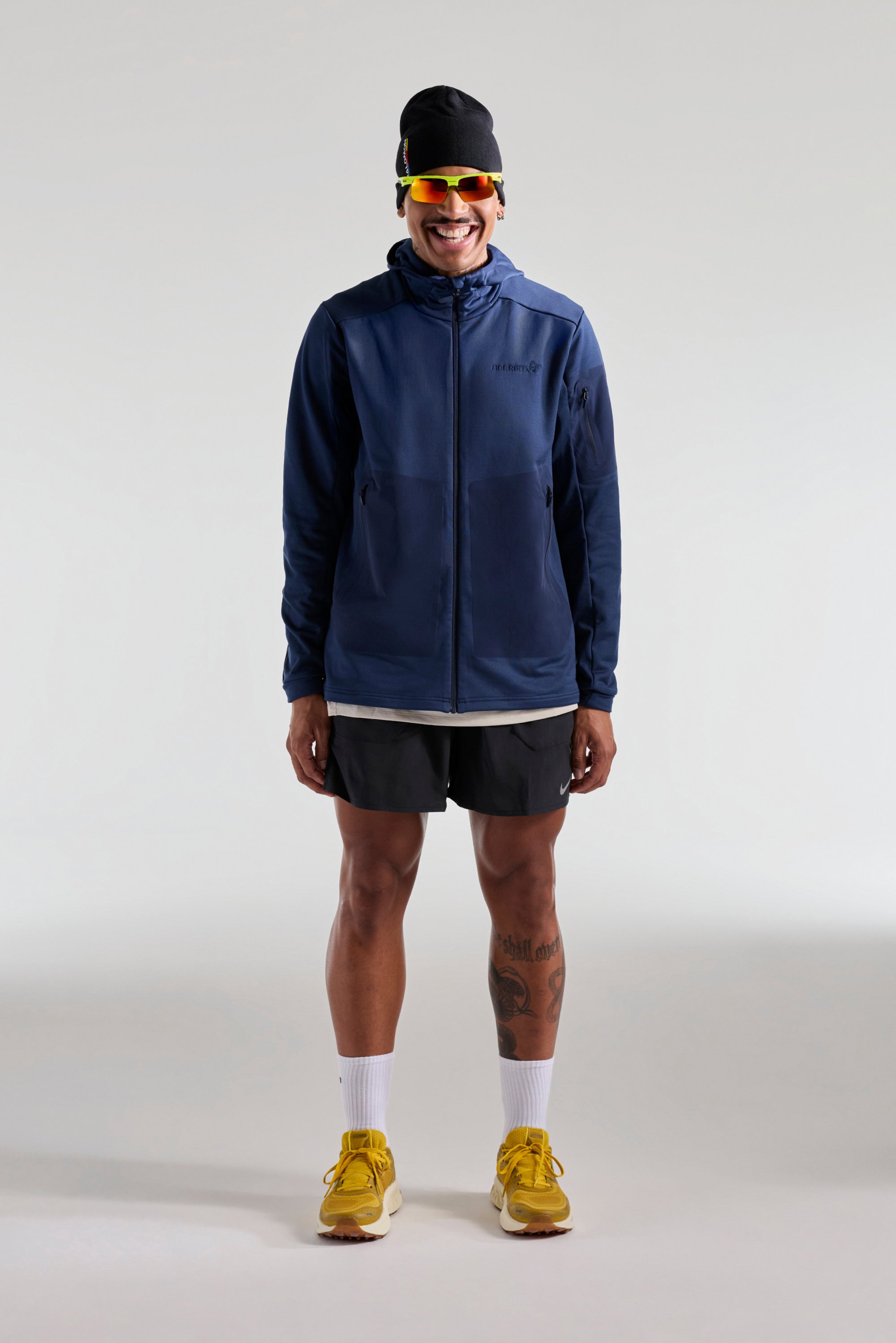 Chaqueta con cremallera en color navy y capucha, con detalles en gris; pantalones cortos negros; zapatillas de running amarillas; calcetines blancos; gafas de sol; diseño minimalista.