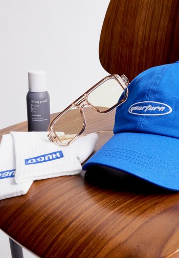 Blaues Cap und weiße Armbänder mit Logos, zusammen mit einer Flasche Trockenshampoo und stilvollen Sonnenbrillen auf einer Holzoberfläche.