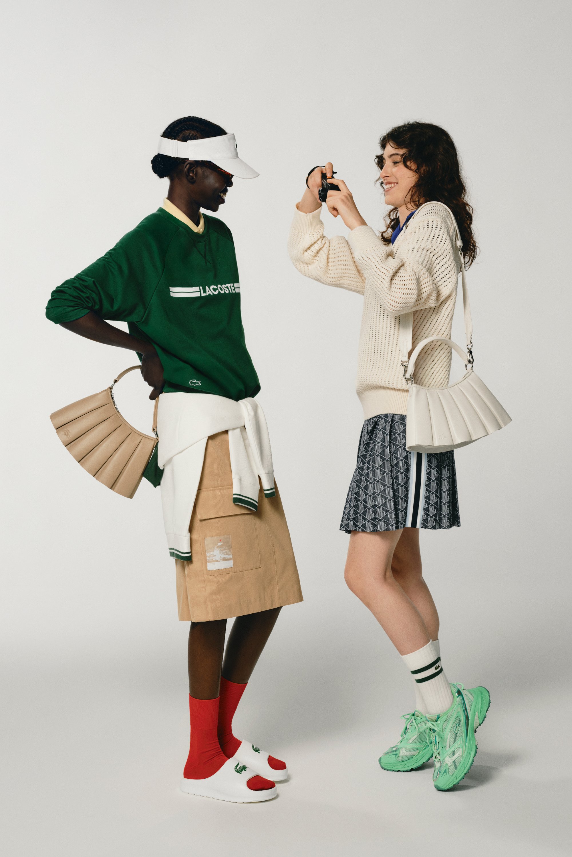 Due donne posano in modo giocoso in outfit alla moda. Una indossa un maglione verde Lacoste e un visore, mentre l'altra sfoggia un maglione di maglia e una gonna a fantasia.