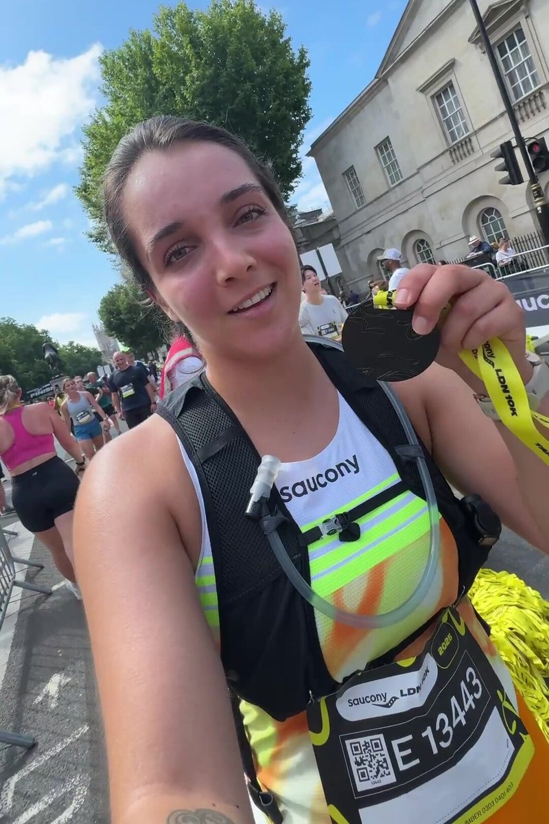 Černá medaile na žluté stuze držená běžcem v tričku Saucony s bílými a fluorescenčními akcenty a hydratačním vpředu.