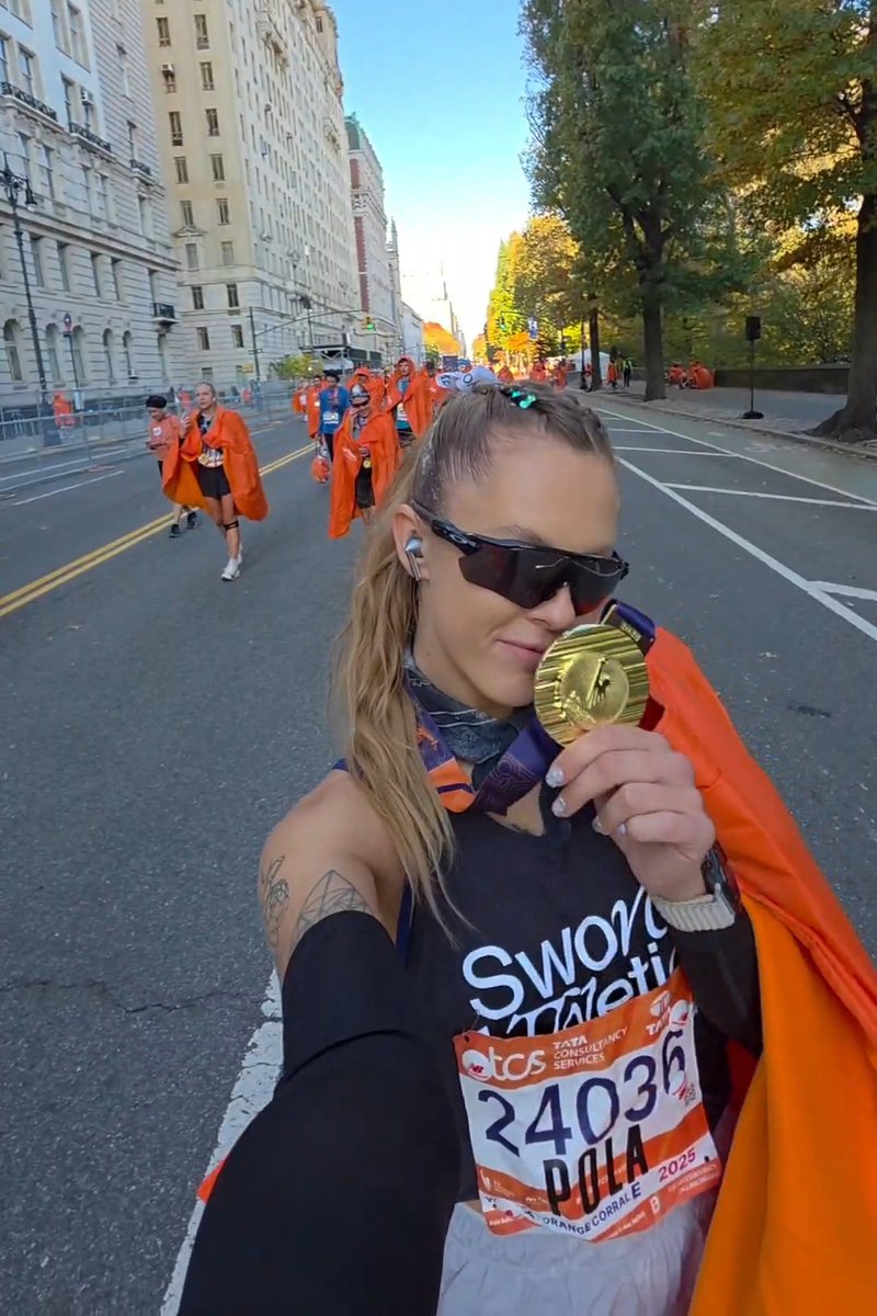 Läufer mit Sonnenbrille und Startnummer 24036 küsst eine Goldmedaille auf einer Straße in der Stadt nach einem Marathon, im Hintergrund sind andere Personen mit orangefarbenen Umhängen zu sehen.
