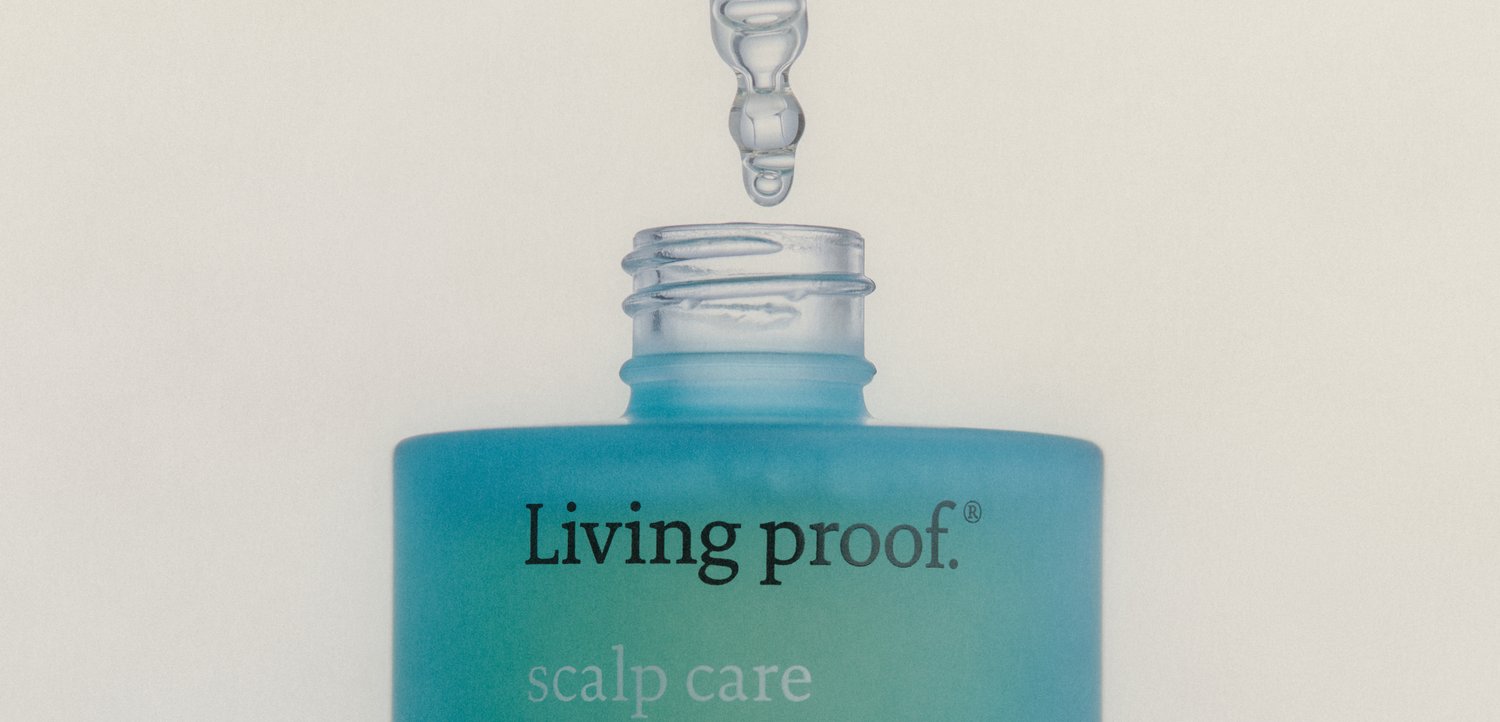 Pipette gibt einen klaren Flüssigkeitstropfen in eine offene blaue Living proof Kopfhautpflegeflasche vor einem einfarbigen Hintergrund ab.