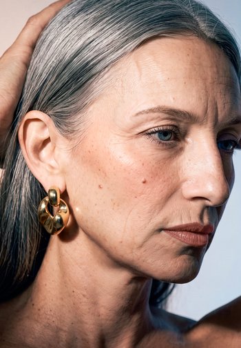Gros plan d'une femme mature aux cheveux gris relevés derrière l'oreille, portant une grande boucle d'oreille en or, regardant sur le côté avec une expression neutre.