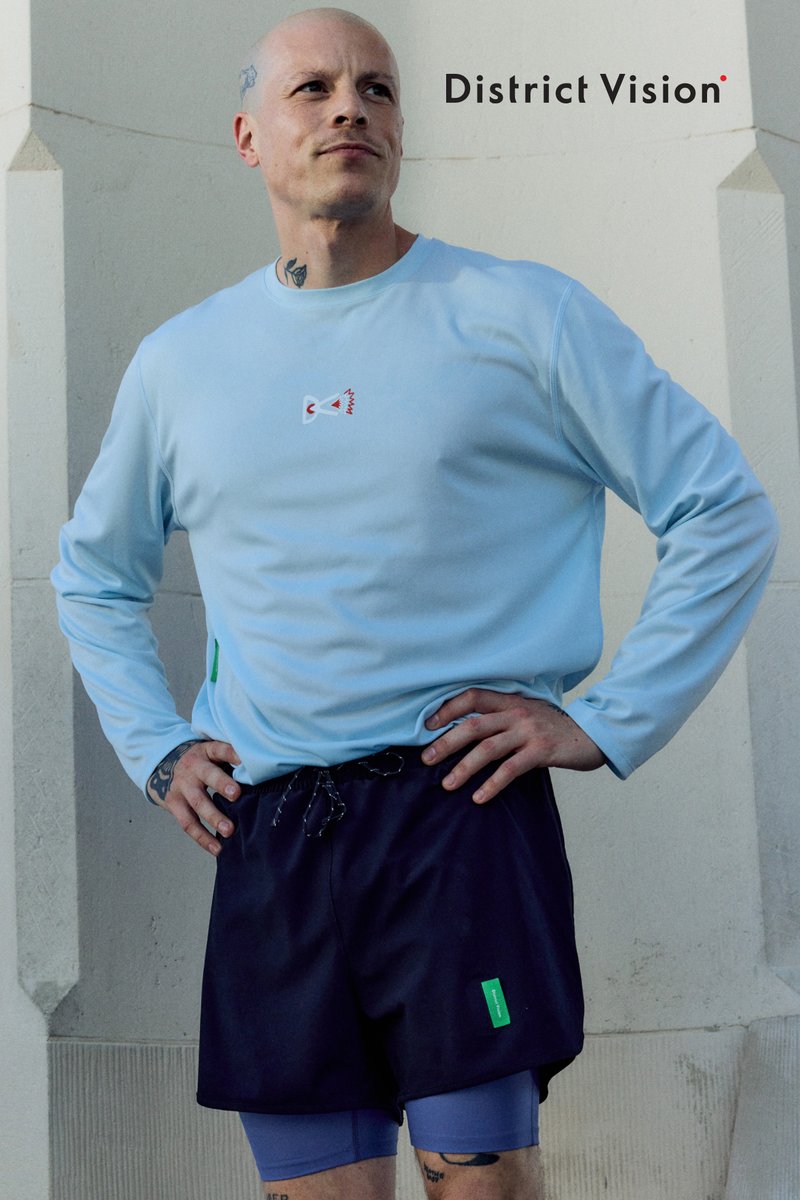 Man with tattoos wearing a light blue long-sleeve shirt and black shorts stands with hands on hips against a pale wall background.