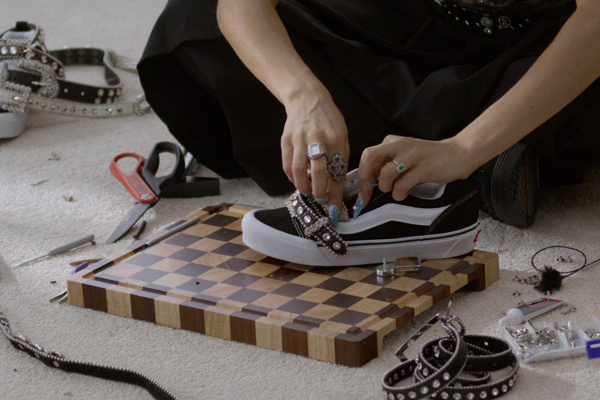 Una persona decora una zapatilla negra con adornos, rodeada de herramientas de manualidades como tijeras y un tablero de ajedrez sobre una alfombra.