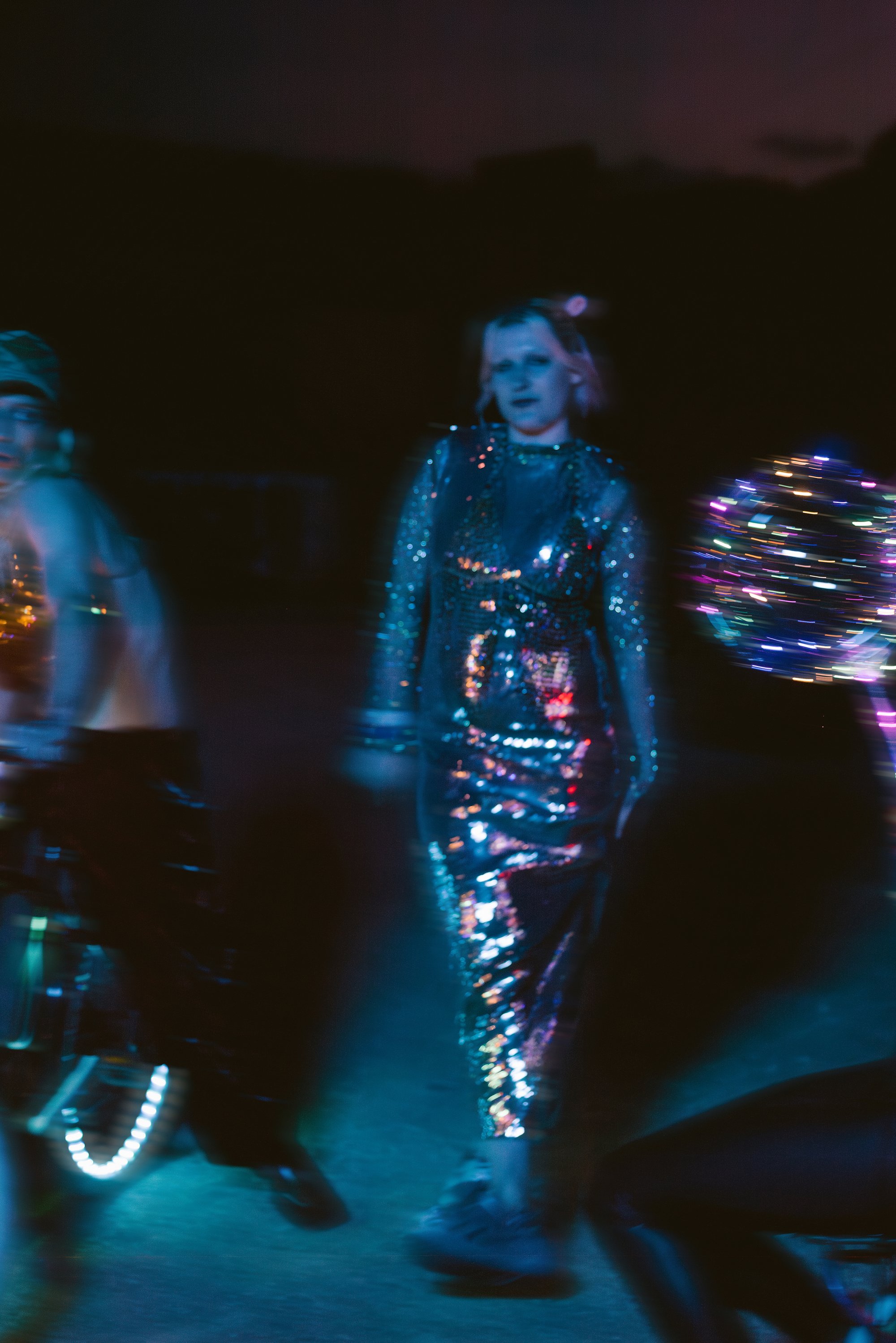 Een groep mensen beweegt energiek in een slecht verlichte ruimte, gehuld in reflecterende, glinsterende outfits met levendige, kleurrijke lichteffecten.