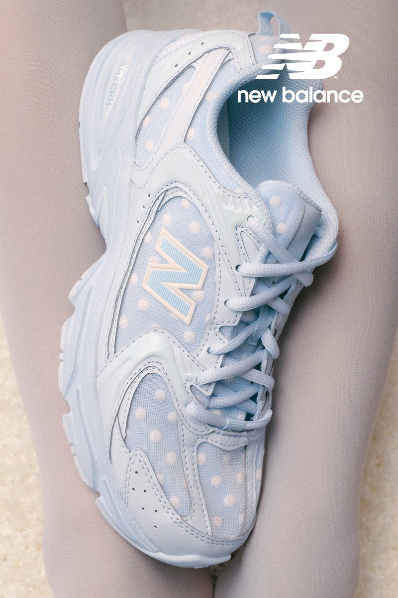 Sneakers azzurri con pois bianchi, caratterizzati da un grande logo "N", poggiati su una superficie morbida. Stile elegante e minimalista.