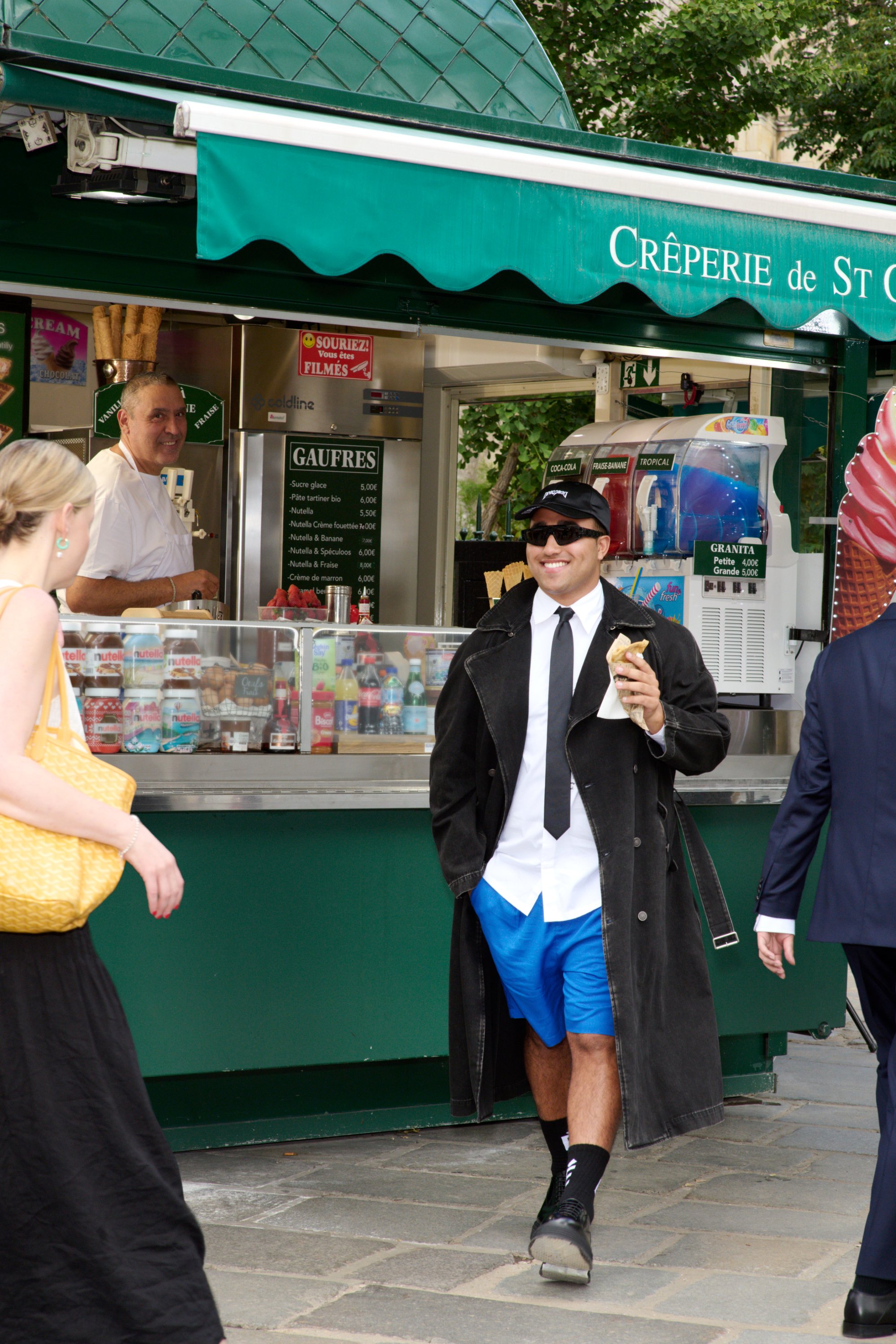 A man in sunglasses, wearing a coat and shorts, smiles while holding food near a crêpe stand, with a chef behind the counter.