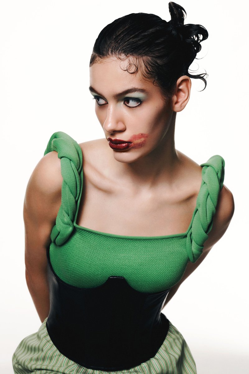 Una modella in un top verde con texture e corsetto nero posa davanti a uno sfondo bianco, presentando un trucco audace con un effetto labbra sfocato.