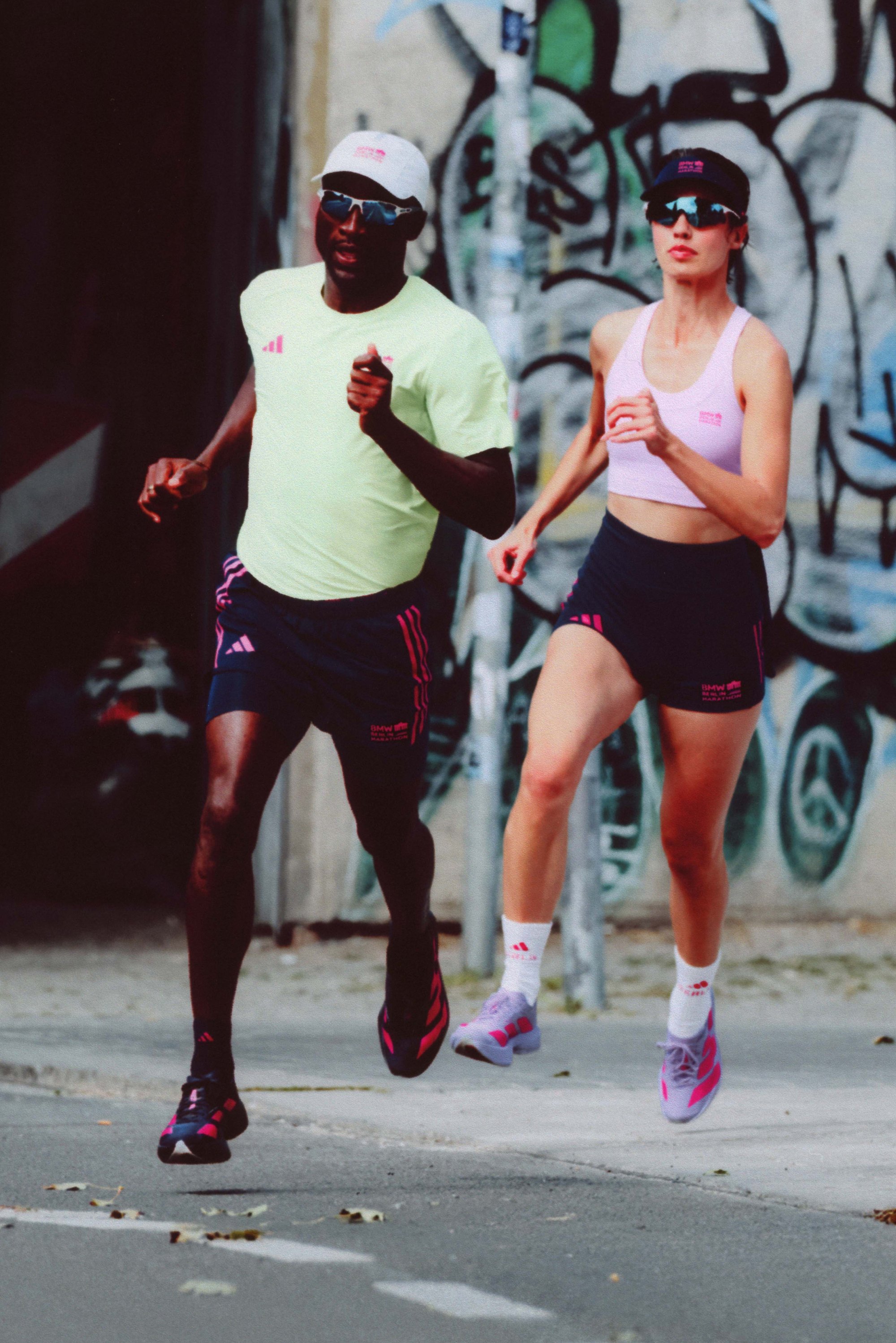Athletic apparel: male in a light green shirt with pink accents, and female in a pink crop top; both wearing dark shorts, running shoes, and caps.