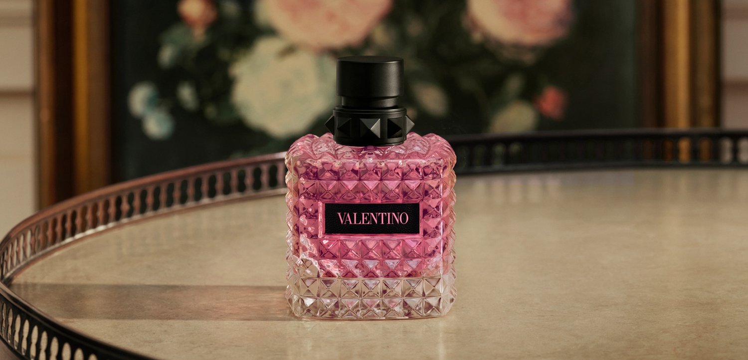 Square faceted glass perfume bottle with pink liquid and black cap labeled Valentino on a round mirrored tray.