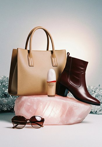 Beżowa skórzana torba, brązowe botki za kostkę na słupku, podkład Shiseido i okulary przeciwsłoneczne w brązowym kolorze w stylu żółwi, ułożone na różowym kryształcie.