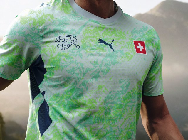 Jasnozielona wzorzysta koszulka sportowa z logo Puma, flagą Szwajcarii i emblematem Szwajcarskiego Związku Piłki Nożnej na piersi, noszona przez osobę na zewnątrz.
