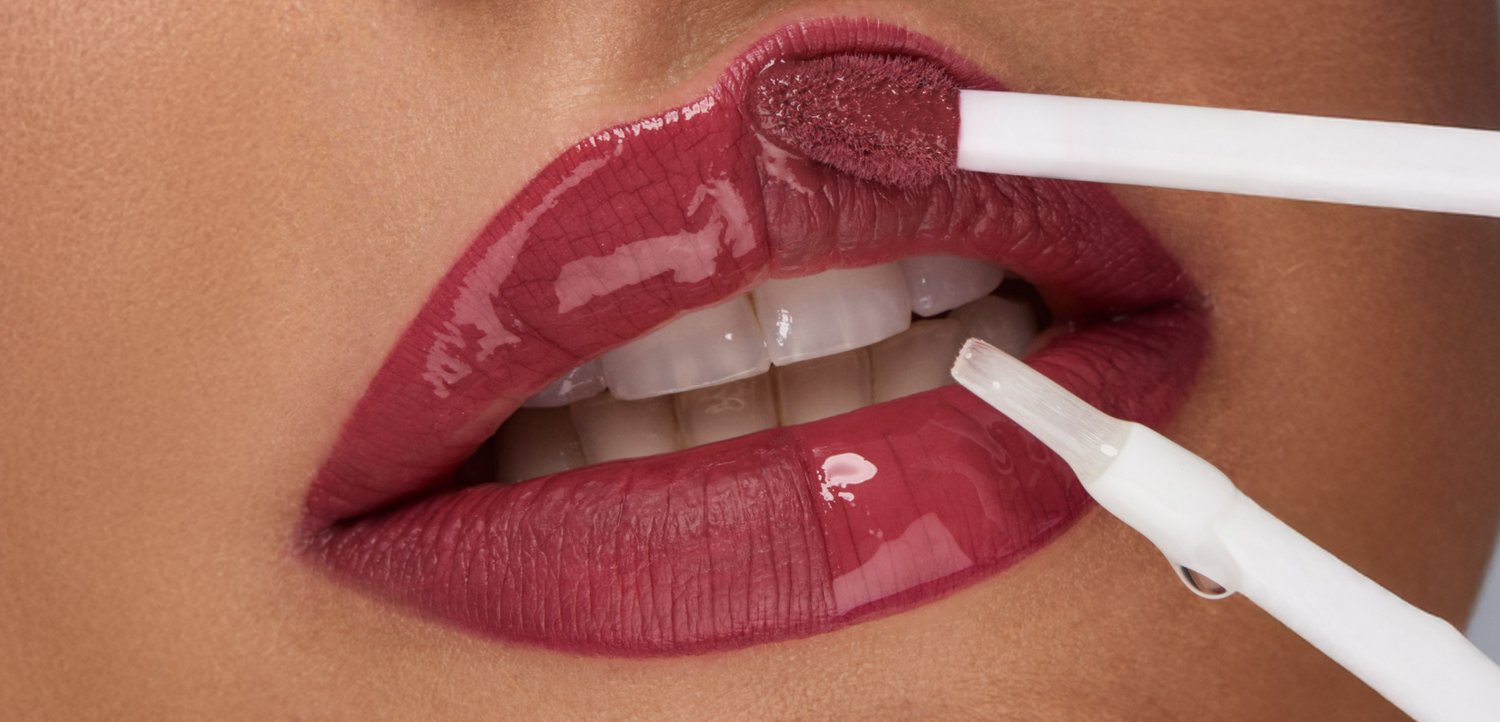 Primer plano de labios aplicando brillo de labios rosa oscuro y brillante con un pincel aplicador mostrado cerca de los labios superiores e inferiores.