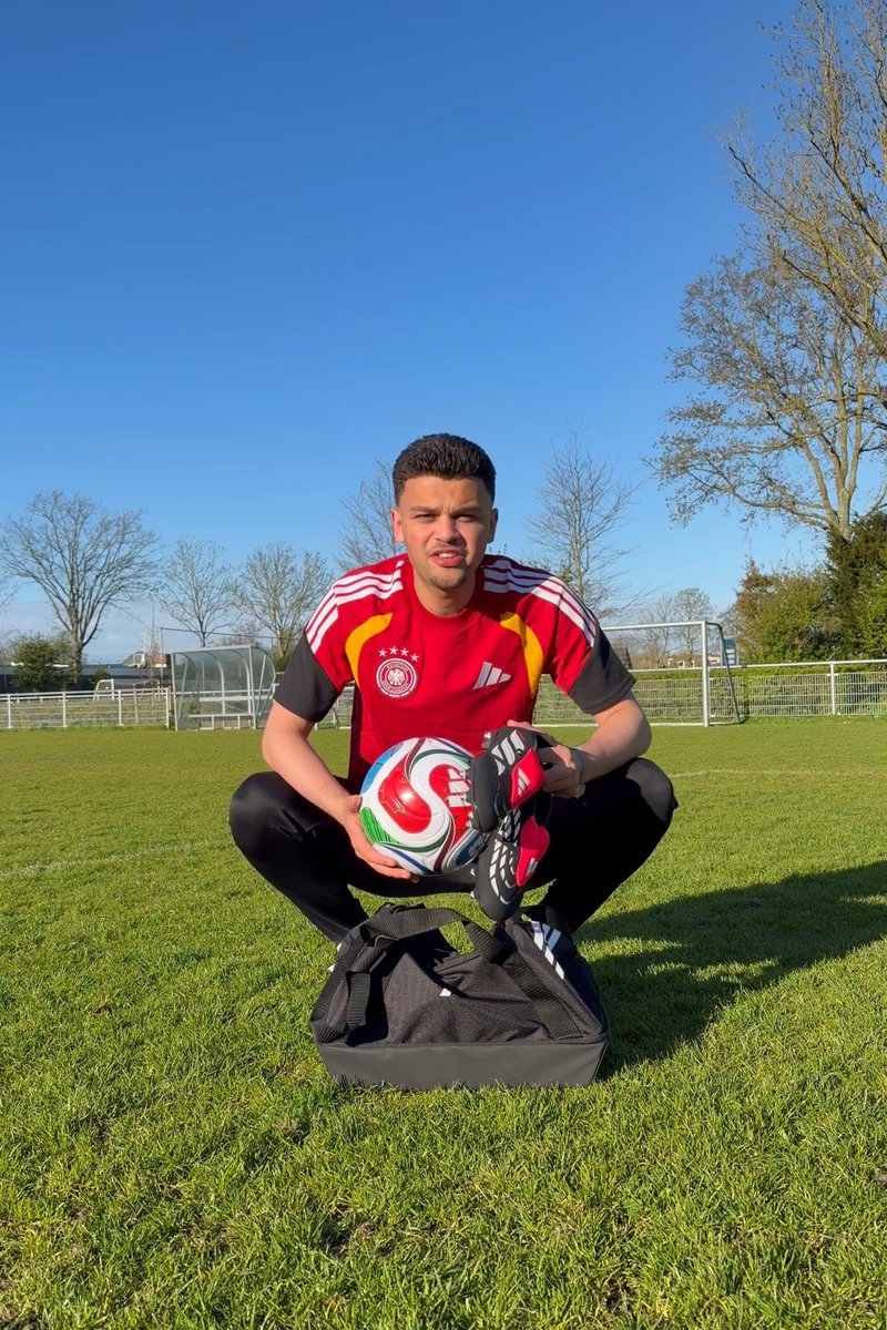 Jonge man hurkt op een grasveld, houdt een voetbal en voetbalschoenen vast, draagt een rood shirt, met een open sporttas voor zich.