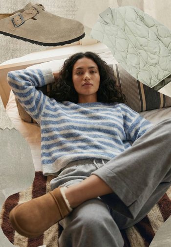 Vrouw die op een bed ligt, gekleed in een blauw gestreepte trui, grijze broek en bruine instapschoenen, met een beige schoen met gesp en een licht gewatteerd jack erboven.