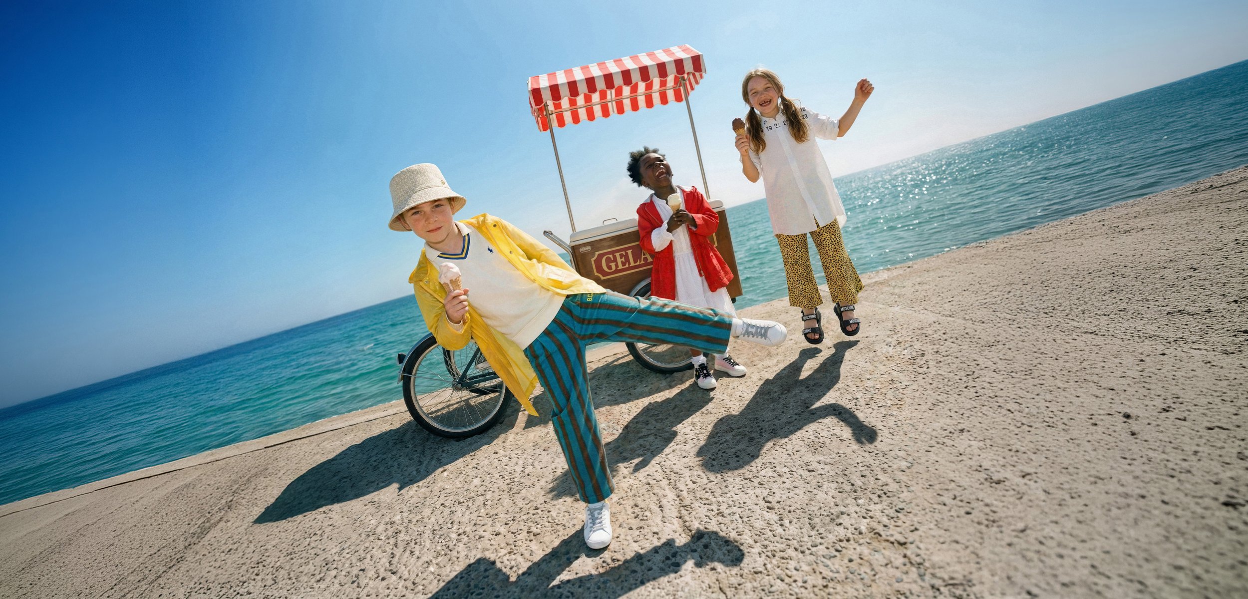 Trois enfants tenant des cornets de glace près d'un chariot à gelato sur une promenade ensoleillée au bord de la mer.
