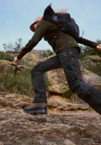 Person wearing hiking gear and backpack climbing rocky terrain under clear sky with sparse vegetation in the background.