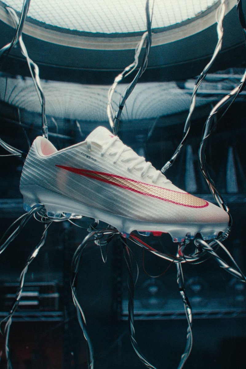 Weißer Nike Fußballschuh mit rotem Swoosh, der an Metallseilen in einer dunklen reflektierenden Umgebung aufgehängt ist.