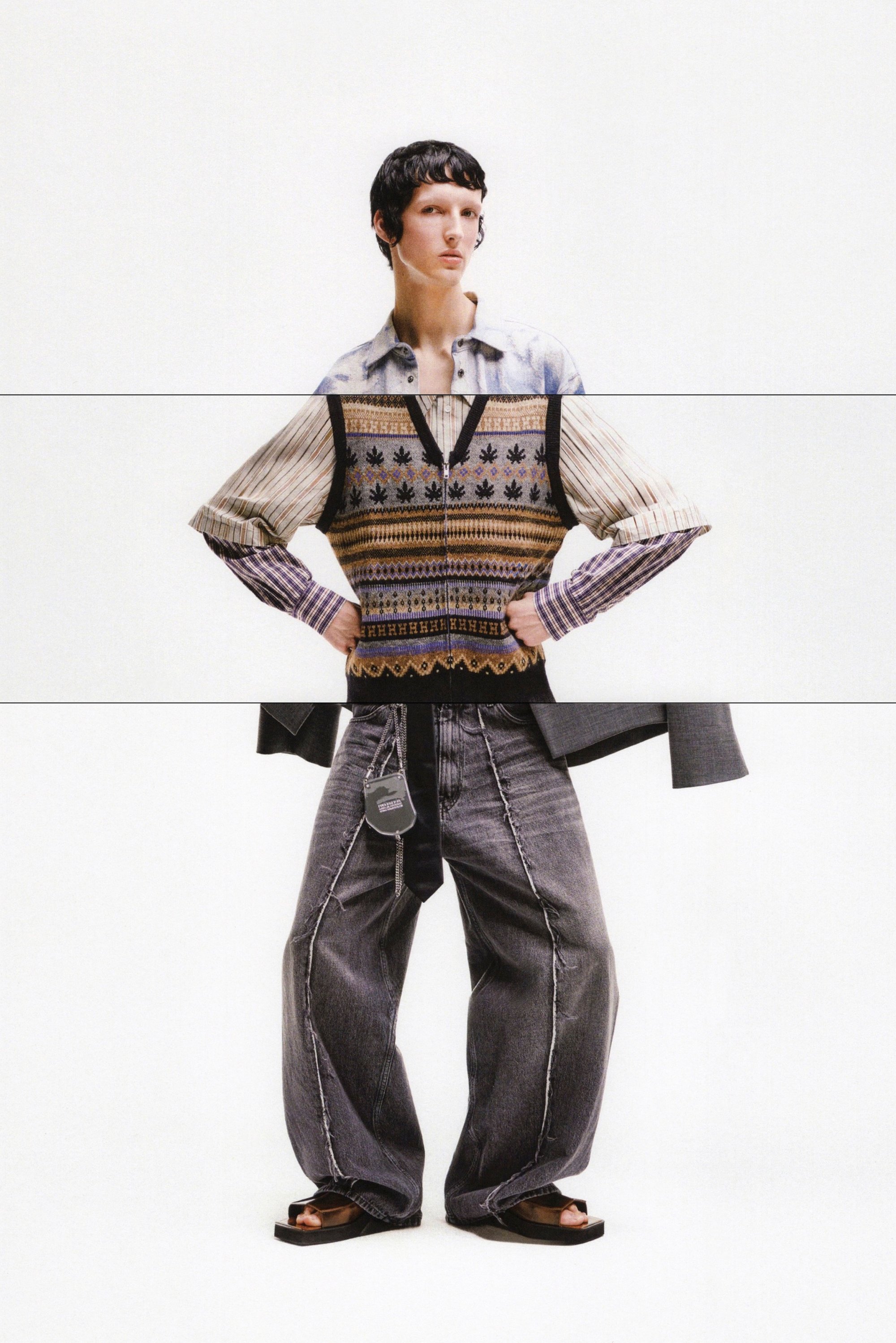 El modelo posa con confianza en un suéter de estampado y jeans grises de gran tamaño, mostrando unas sandalias elegantes frente a un fondo blanco liso.