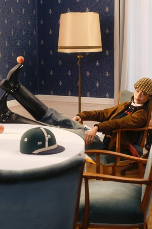 A casually dressed person reclines in a chair, feet on a table with an apple and a cap nearby, framed by a patterned wall and soft lighting.
