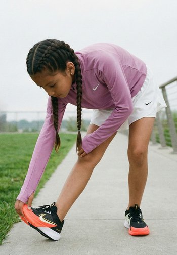 Junges Mädchen mit geflochtenen Haaren, das eine lila Jacke und weiße Shorts trägt, dehnt ihr Bein auf einem Weg in der Nähe von grünem Gras und einem Geländer im Freien.